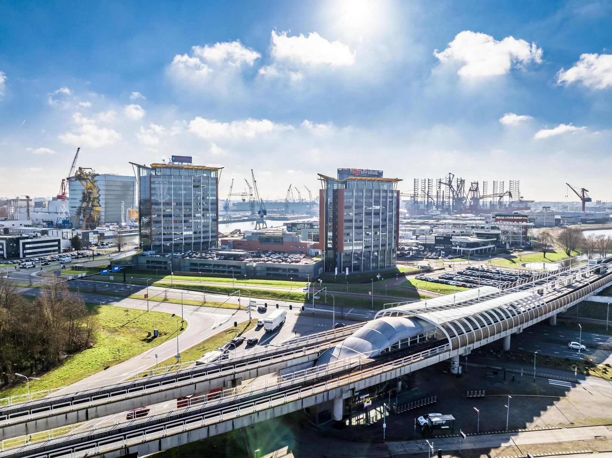 Luchtfoto van de Karel Doormanweg in Rotterdam met kantoorgebouwen, metrostation en havenkranen op de achtergrond.
